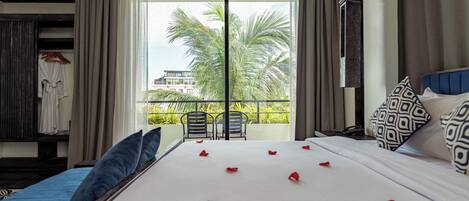 A hotel room with a large bed, a balcony with chairs, and a view of palm trees.
