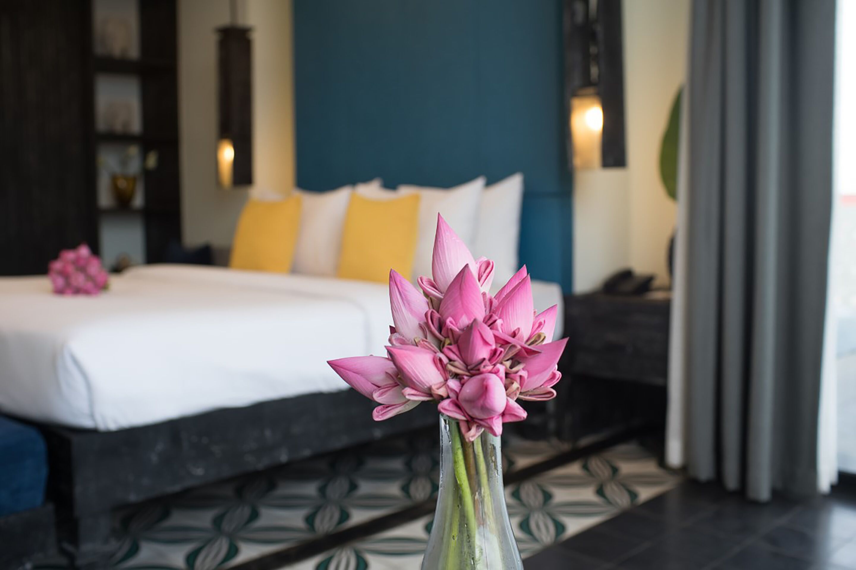Una cama con ropa de cama blanca, un florero con flores rosas y un suelo estampado.