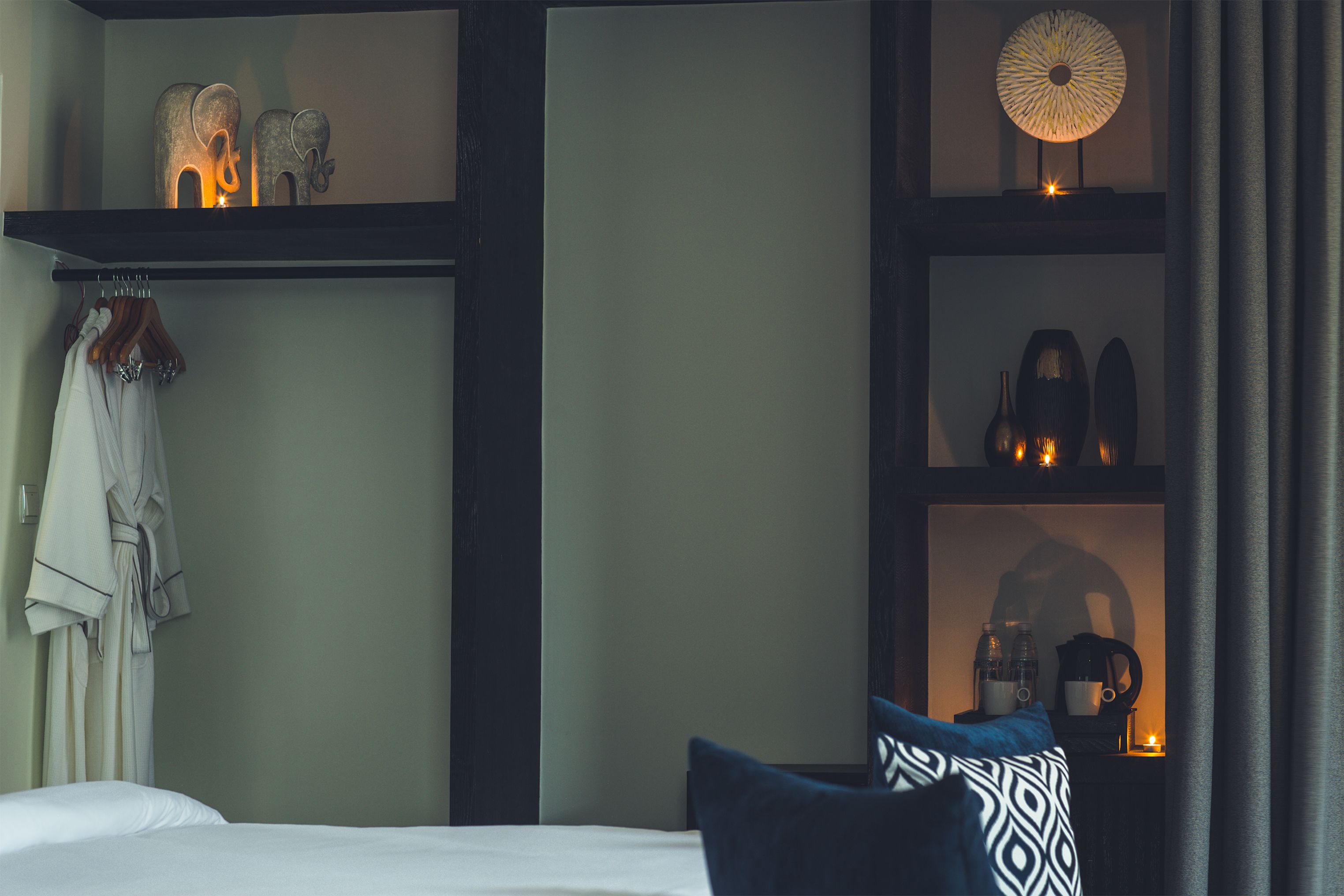 Een hotelkamer met een bed, een badjas hangend aan de muur, een plank met decoratieve objecten, en een nachtkastje met een lamp en een beker.