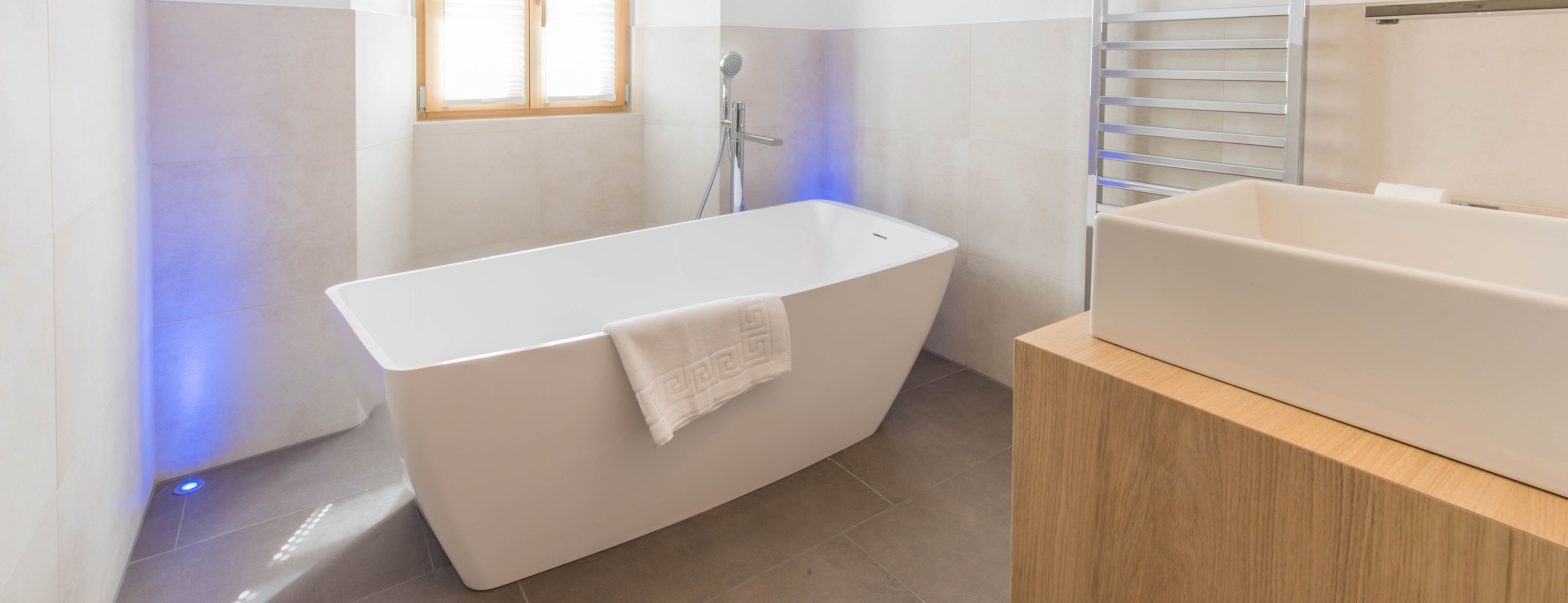 Een moderne badkamer met een vrijstaand bad, een houten wastafelmeubel en een handdoek aan het bad.
