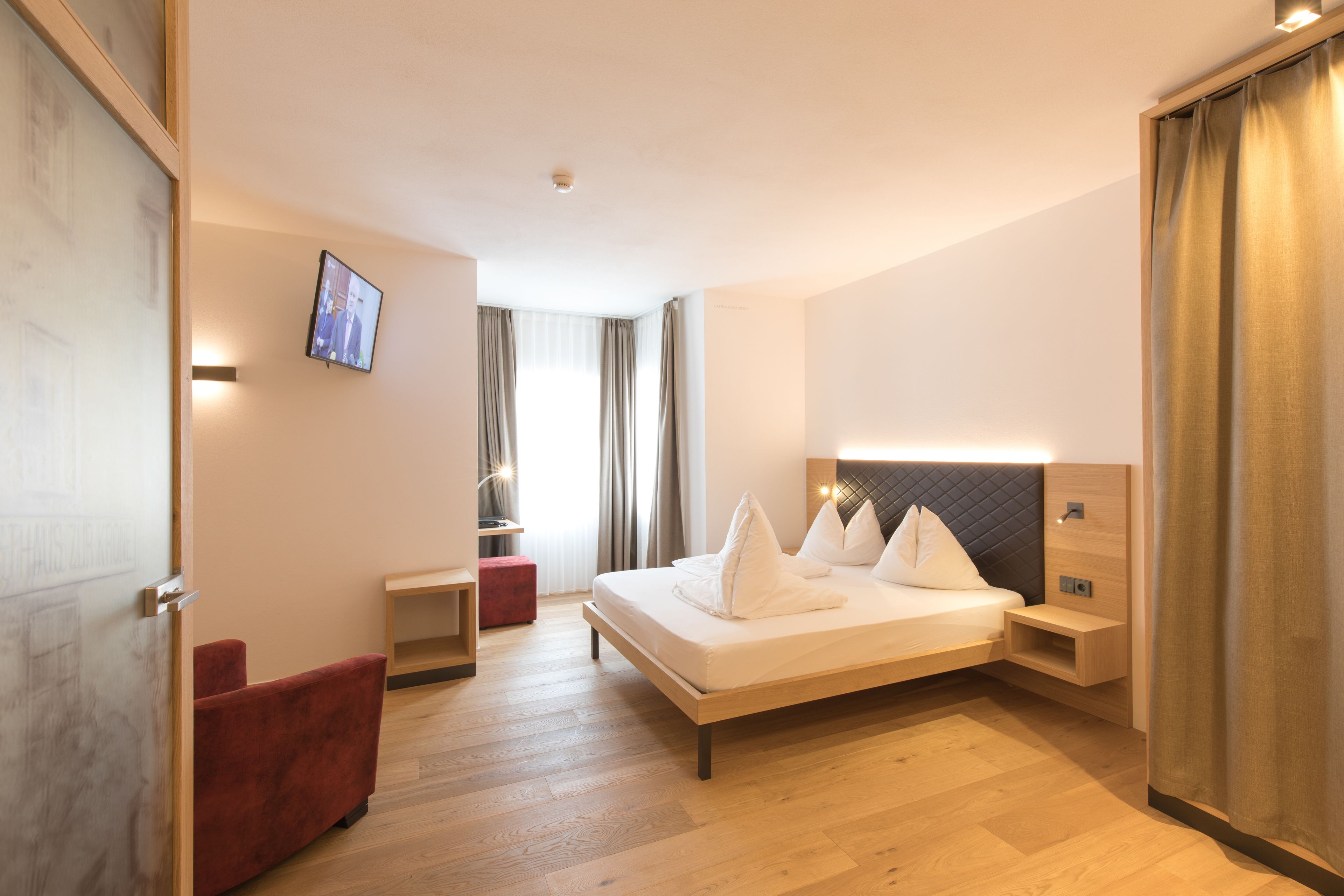 A modern hotel room with a large bed, a desk, a red chair, and a TV on the wall.