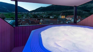 A hot tub in a room with a view of a village and mountains.