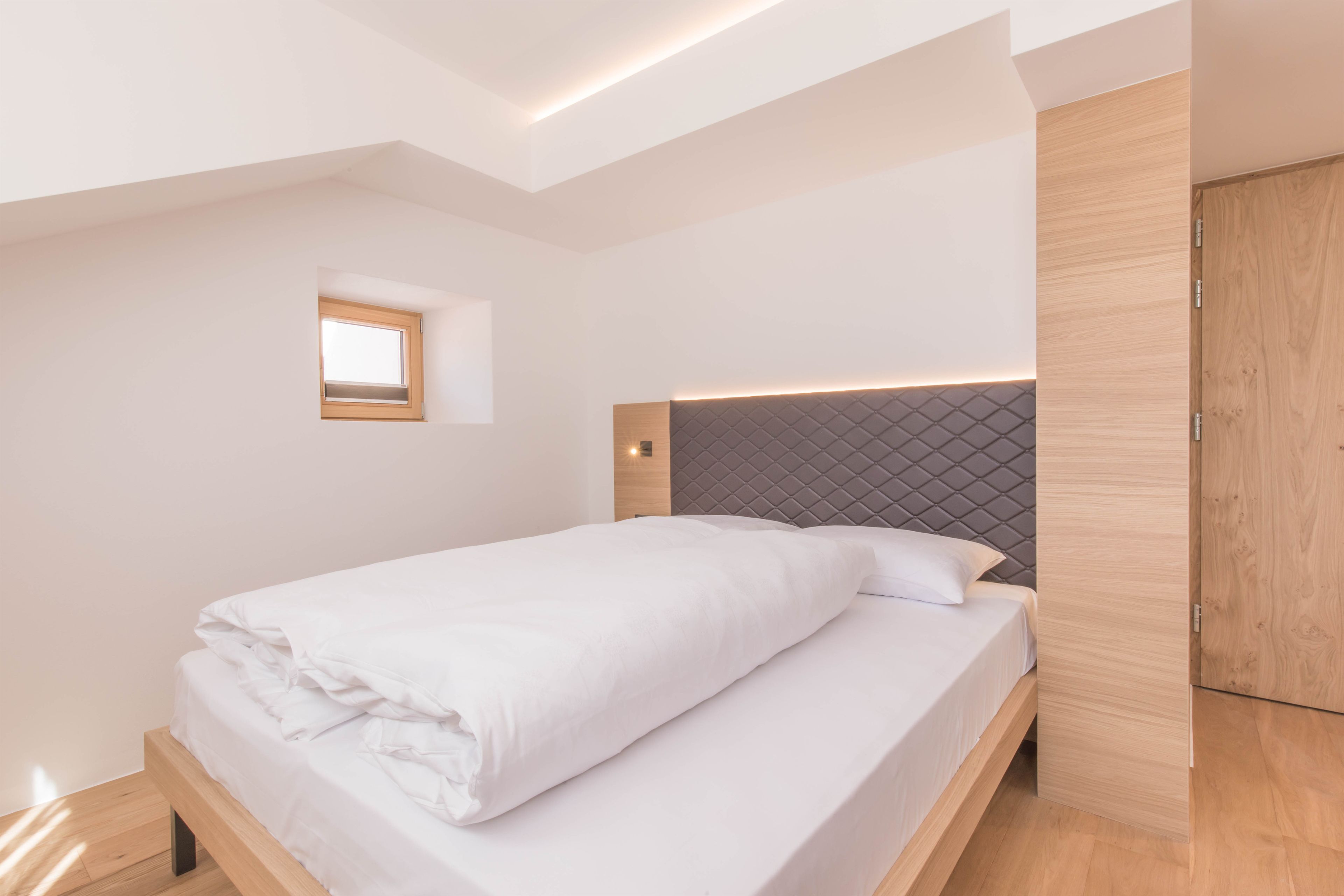 Ein modernes Schlafzimmer mit einem Bett, einem kleinen Fenster und einem Holzschrank.