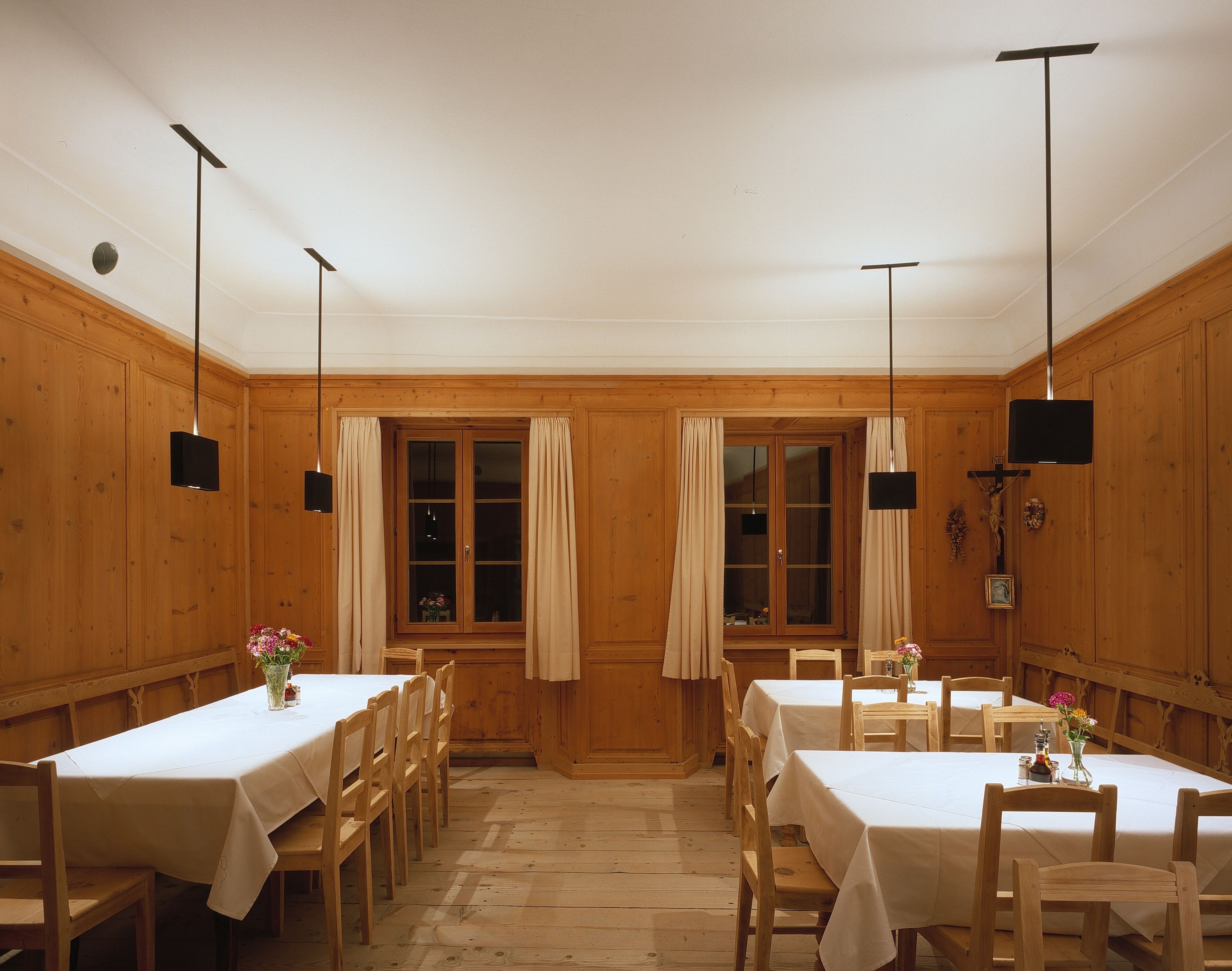 A dining area with wooden walls and tables, featuring a central window with curtains, pendant lights, and a crucifix on the wall.