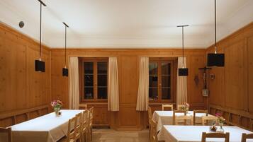 A dining area with wooden walls and tables, featuring a central window with curtains, pendant lights, and a crucifix on the wall.