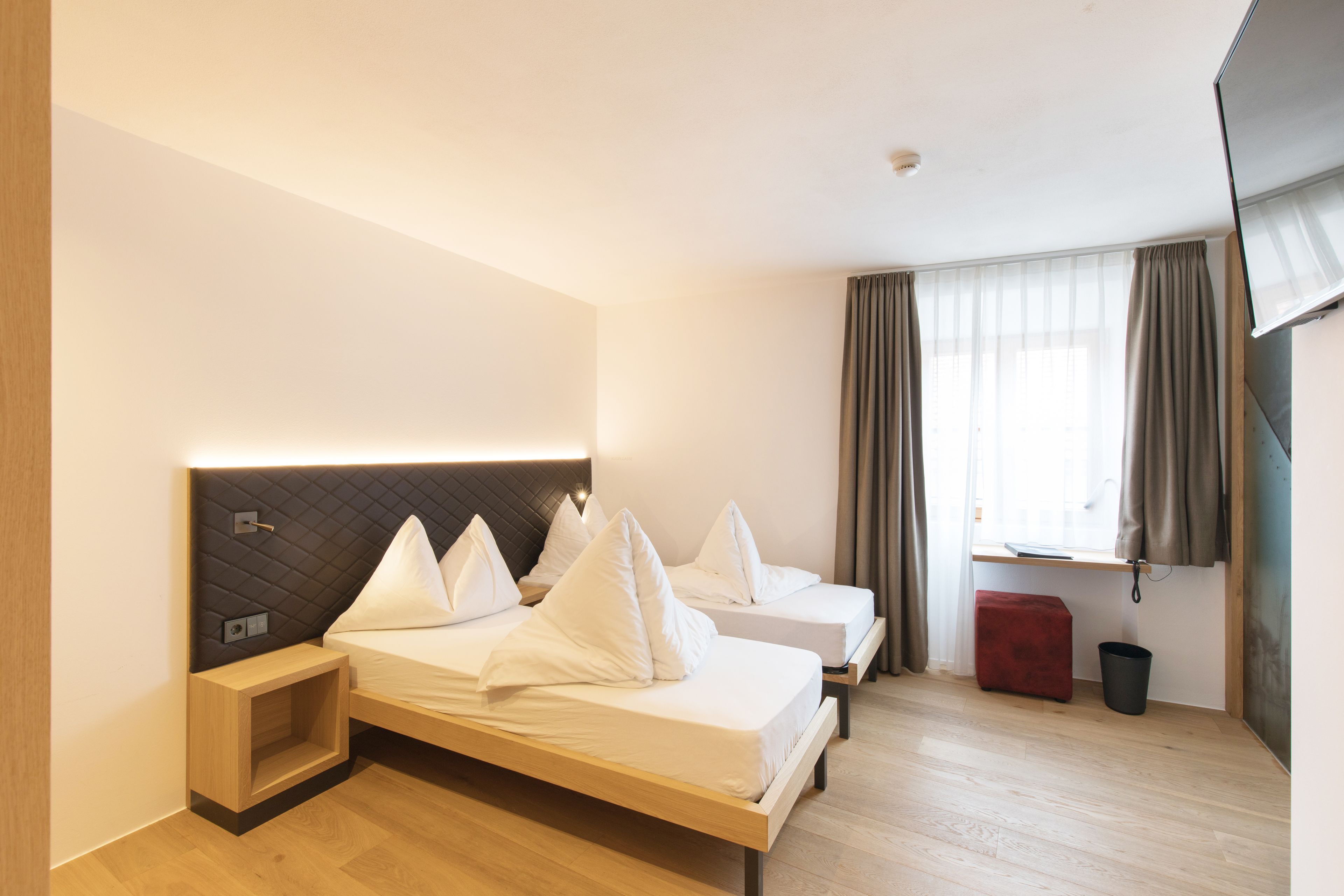 Una habitación de hotel moderna con dos camas, una mesita de noche, un taburete rojo y una ventana con cortinas.