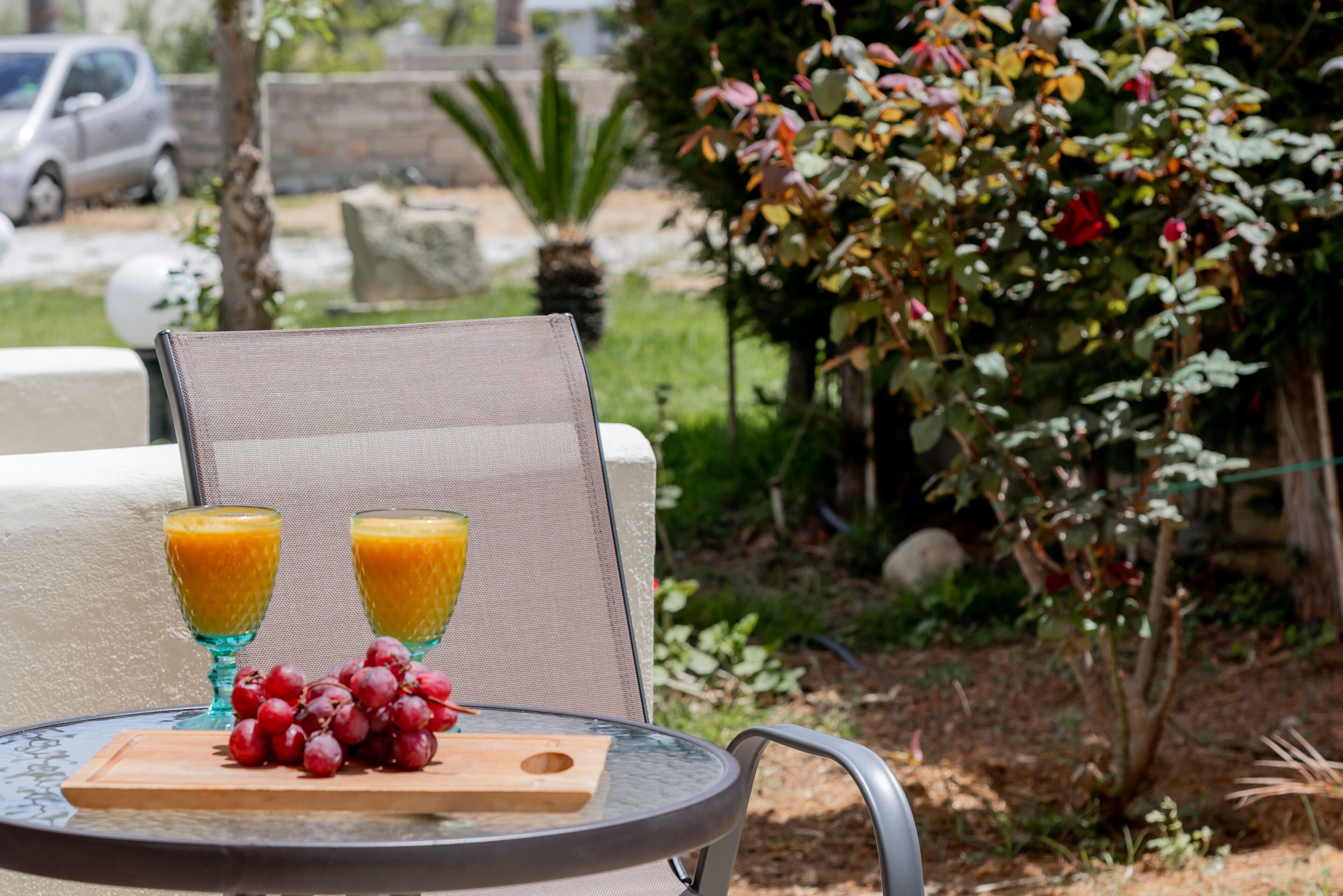 Une table en verre avec deux verres de jus d’orange, une planche en bois avec des raisins rouges et une chaise avec un dossier en treillis.