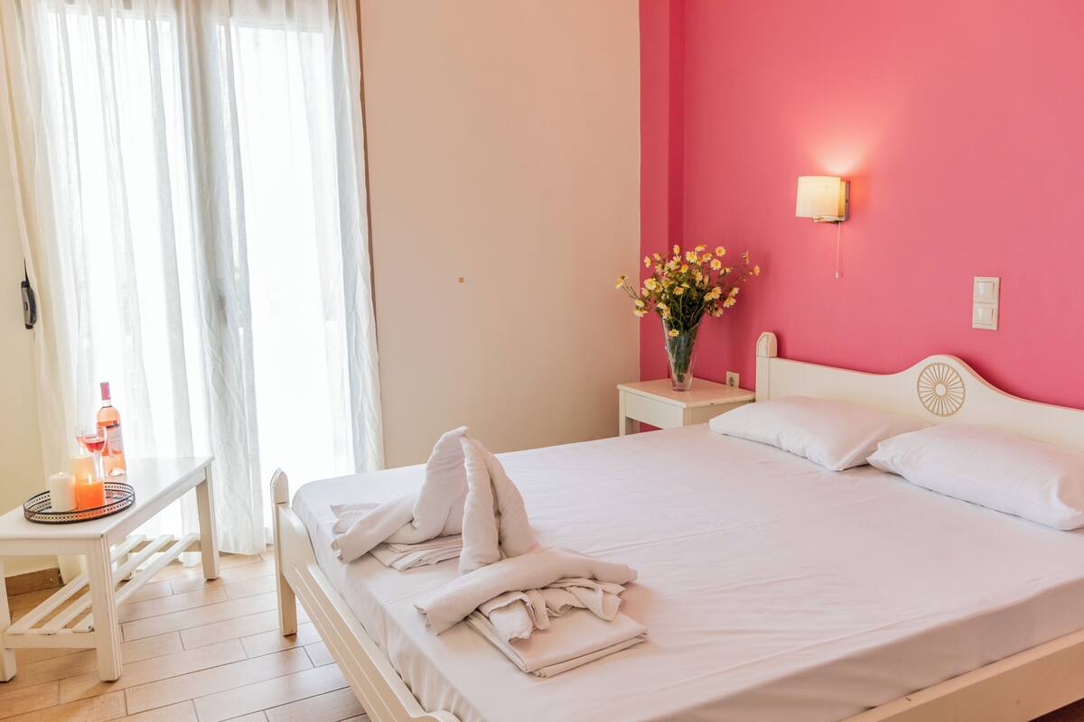 A neatly made bed with pillows and folded towels, a bedside table with a vase of flowers, and a window with sheer curtains.