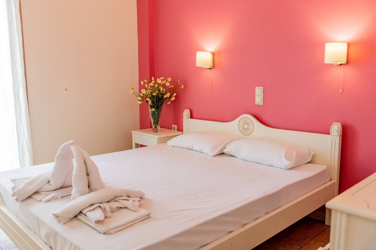 A neatly made bed with white linens and towels, a vase of flowers on a bedside table, and a wall-mounted light fixture.
