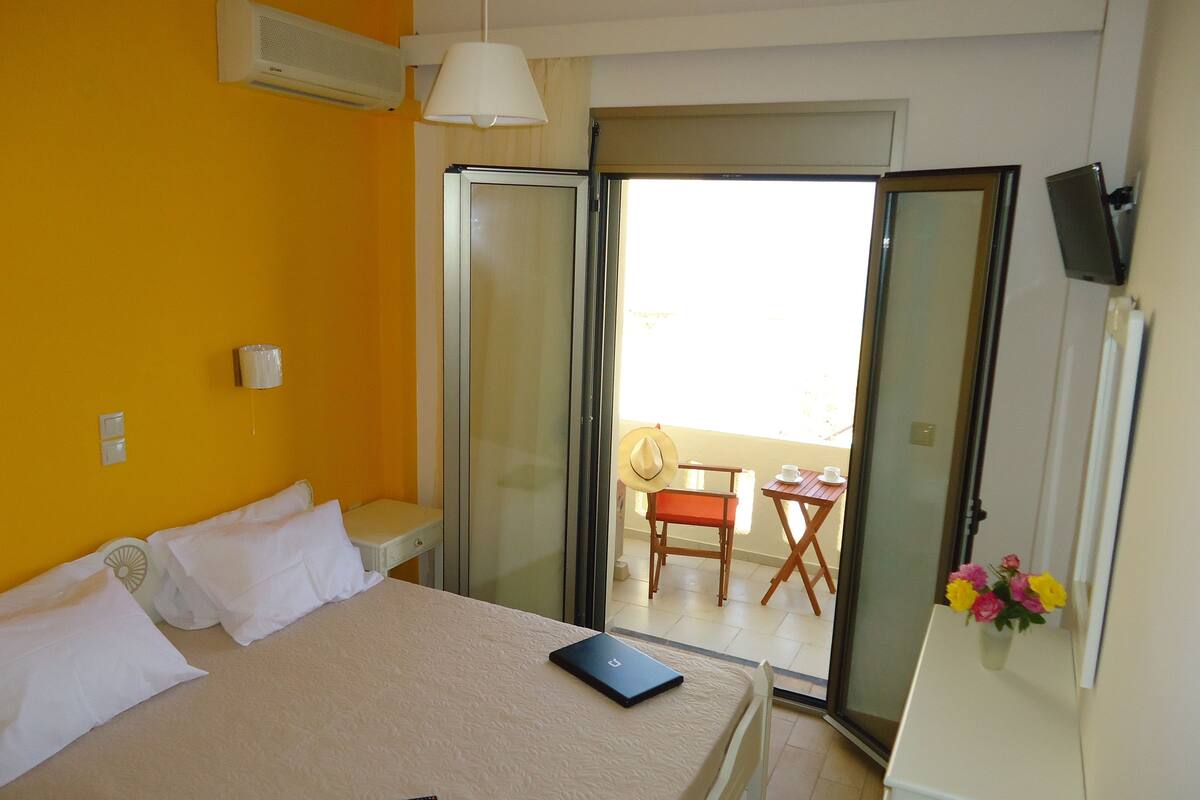 A hotel room with a bed, a remote control, a laptop, a sliding glass door leading to a balcony with a chair and table, and a vase of flowers.