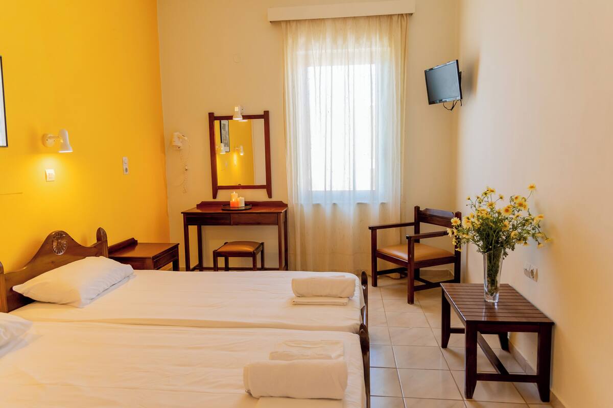 A hotel room with two beds, a desk, a chair, a TV, and a vase with flowers.