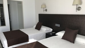 Habitación de hotel con dos camas, cada una con ropa blanca y mantas marrones. Hay dos lámparas fijadas a la pared y una mesita de noche pequeña con un control remoto.