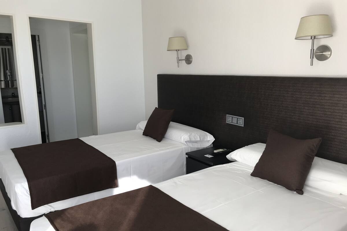 A hotel room with two beds, each with white linens and brown throw blankets. There are two wall-mounted lamps and a small bedside table with a remote control.