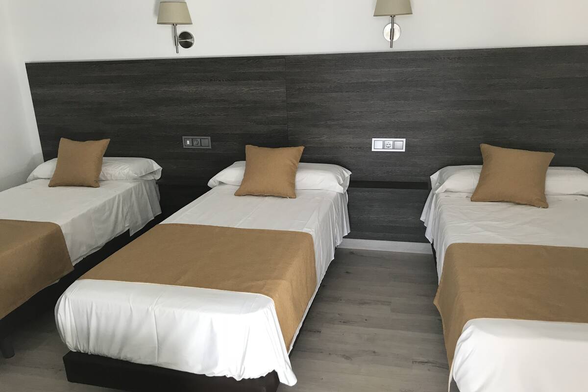 A hotel room with four single beds, each with a brown pillow and white bedding. There are two wall-mounted lamps on either side of the room.