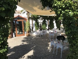 Un área de estar al aire libre con sillas y mesas de plástico blanco, una pérgola de madera y plantas en macetas.