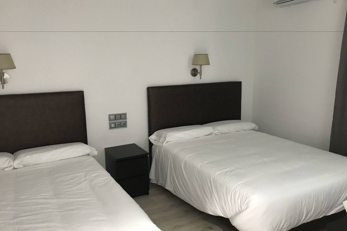 A hotel room with two single beds, a nightstand, a wall socket, and a wall-mounted air conditioning unit.
