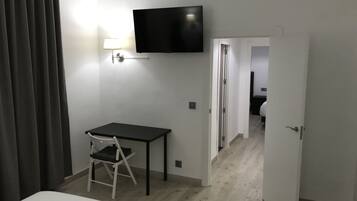Habitación de hotel con una cama, un escritorio con silla, un televisor montado en la pared y una puerta que da a otra habitación.