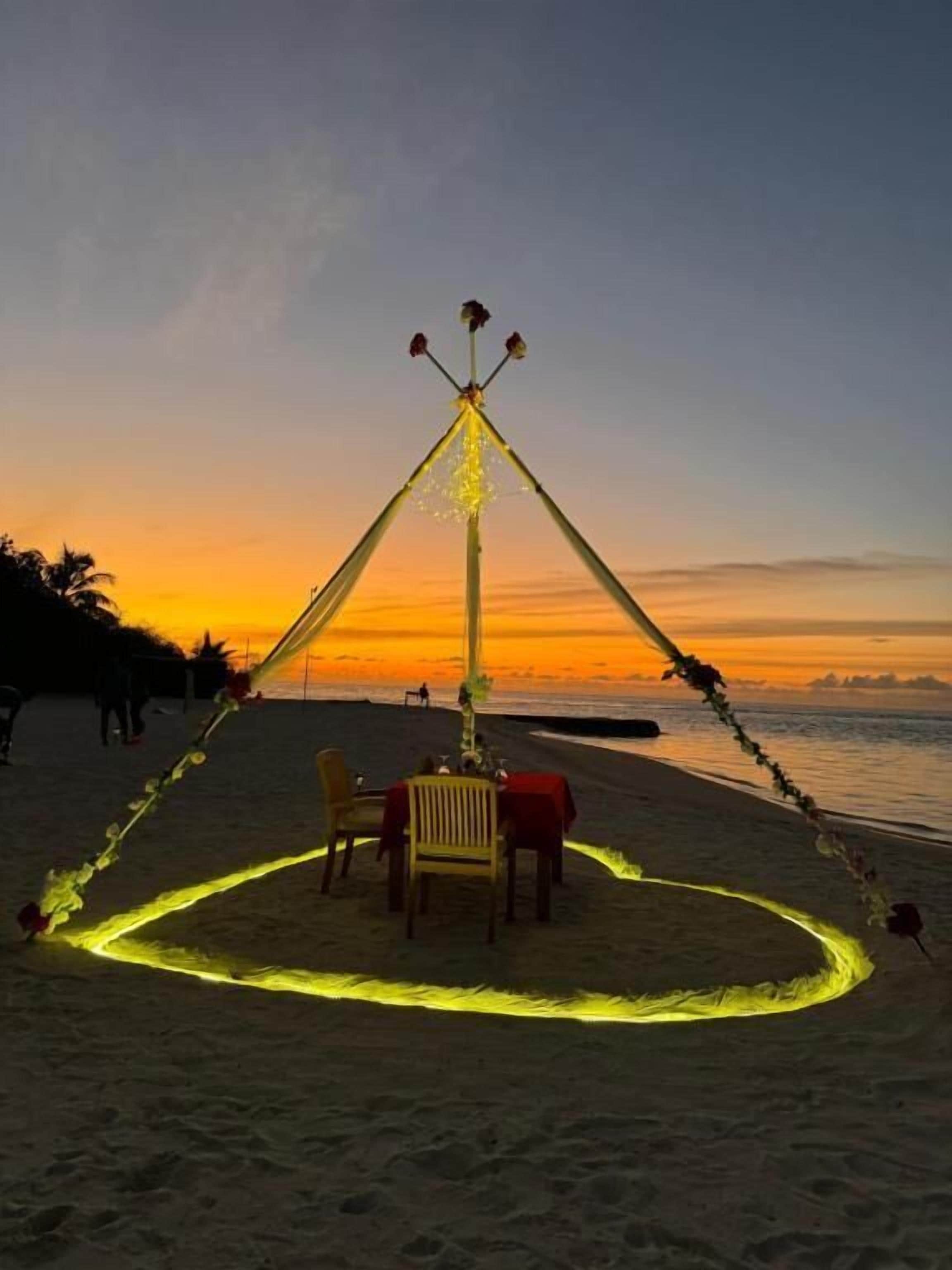 Un décor de plage avec une table et des chaises disposées pour un repas, le tout entouré d’un chemin illuminé à des fins décoratives.