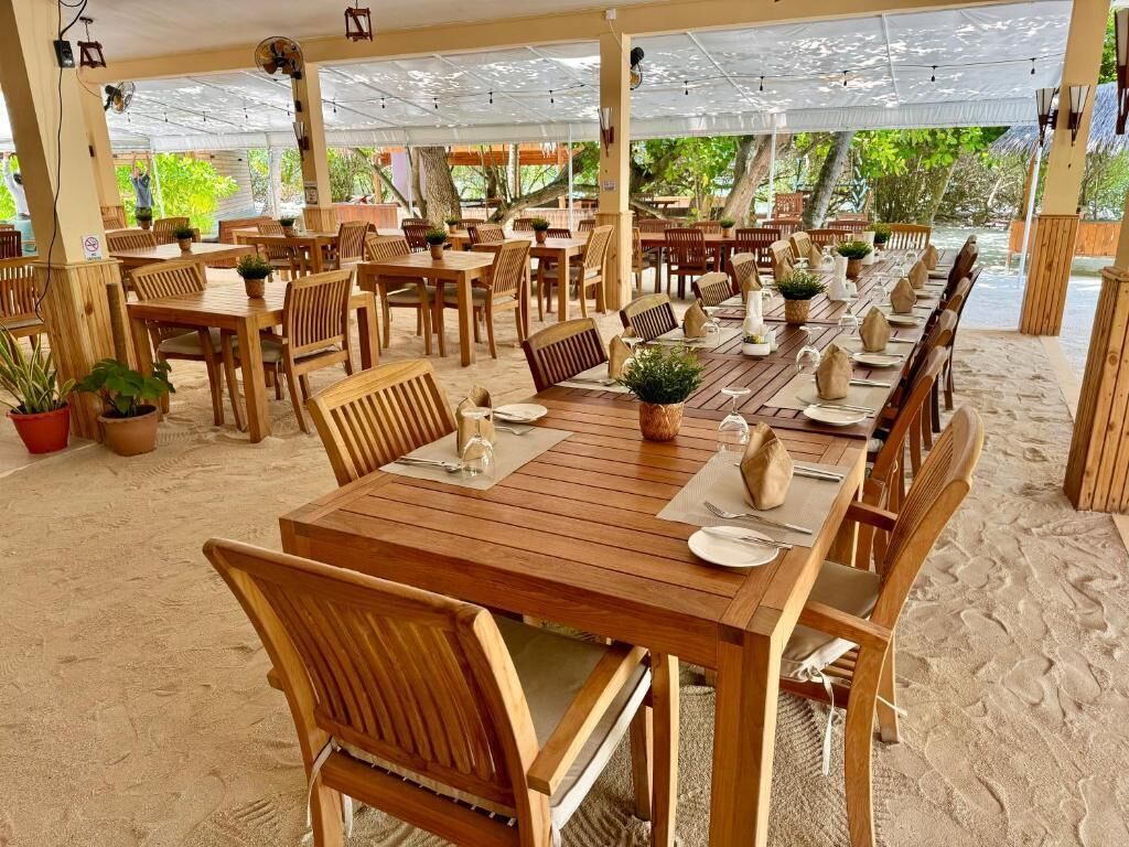 An outdoor dining area with wooden tables and chairs, surrounded by potted plants and a sandy floor.
