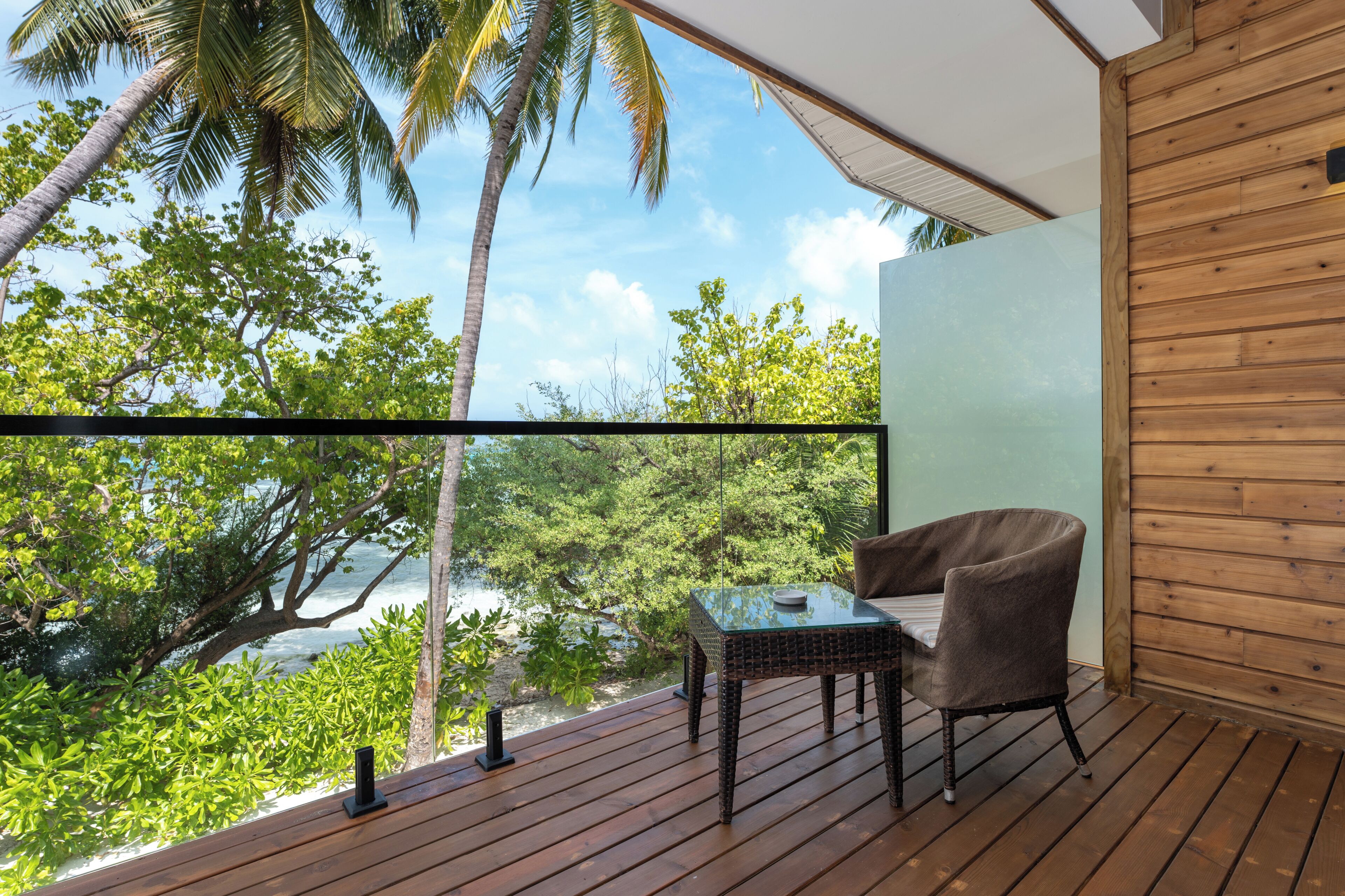 A balcony with a glass table and chair, overlooking a tropical landscape with palm trees and a clear sky.