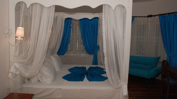 Un llit amb cortines i draperiades blanques lleugeres, accents blaus, una làmpada de nit i una cadira color aguamarina en una habitació amb terres de fusta.