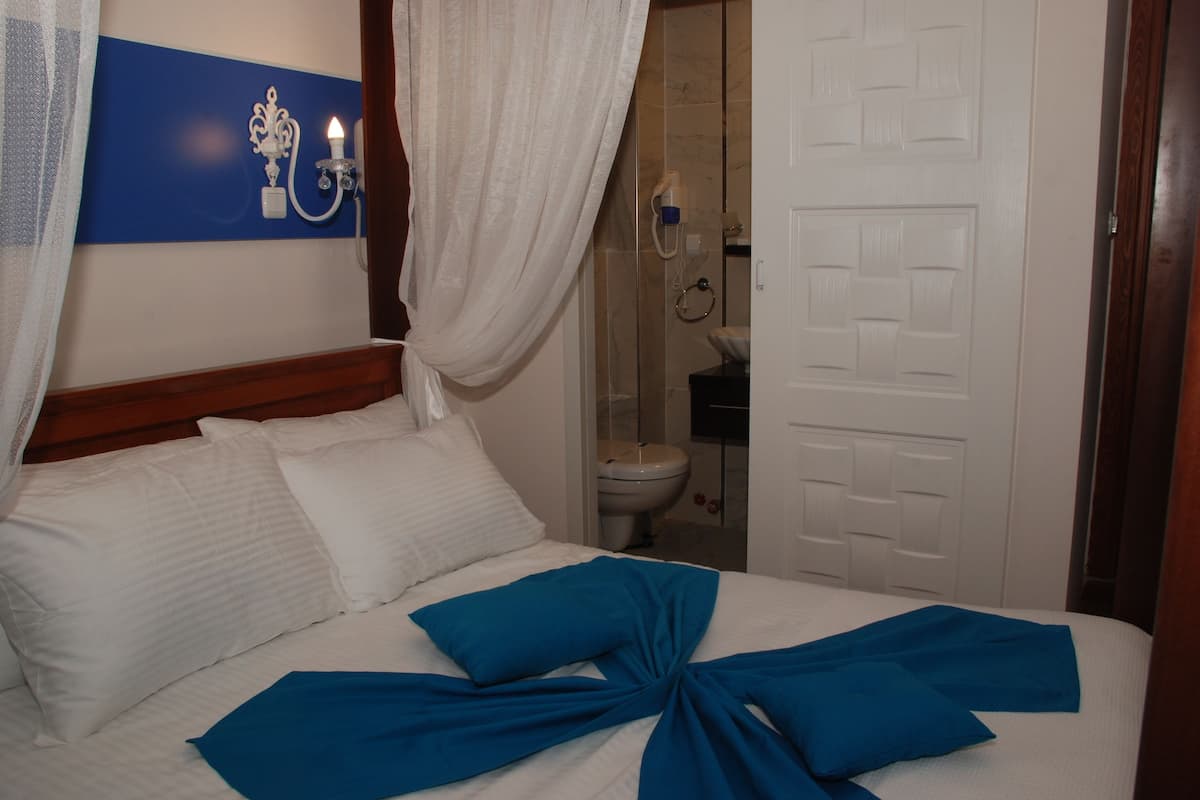 A hotel room with a bed, white curtains, a blue wall decoration, and a bathroom visible through an open door.