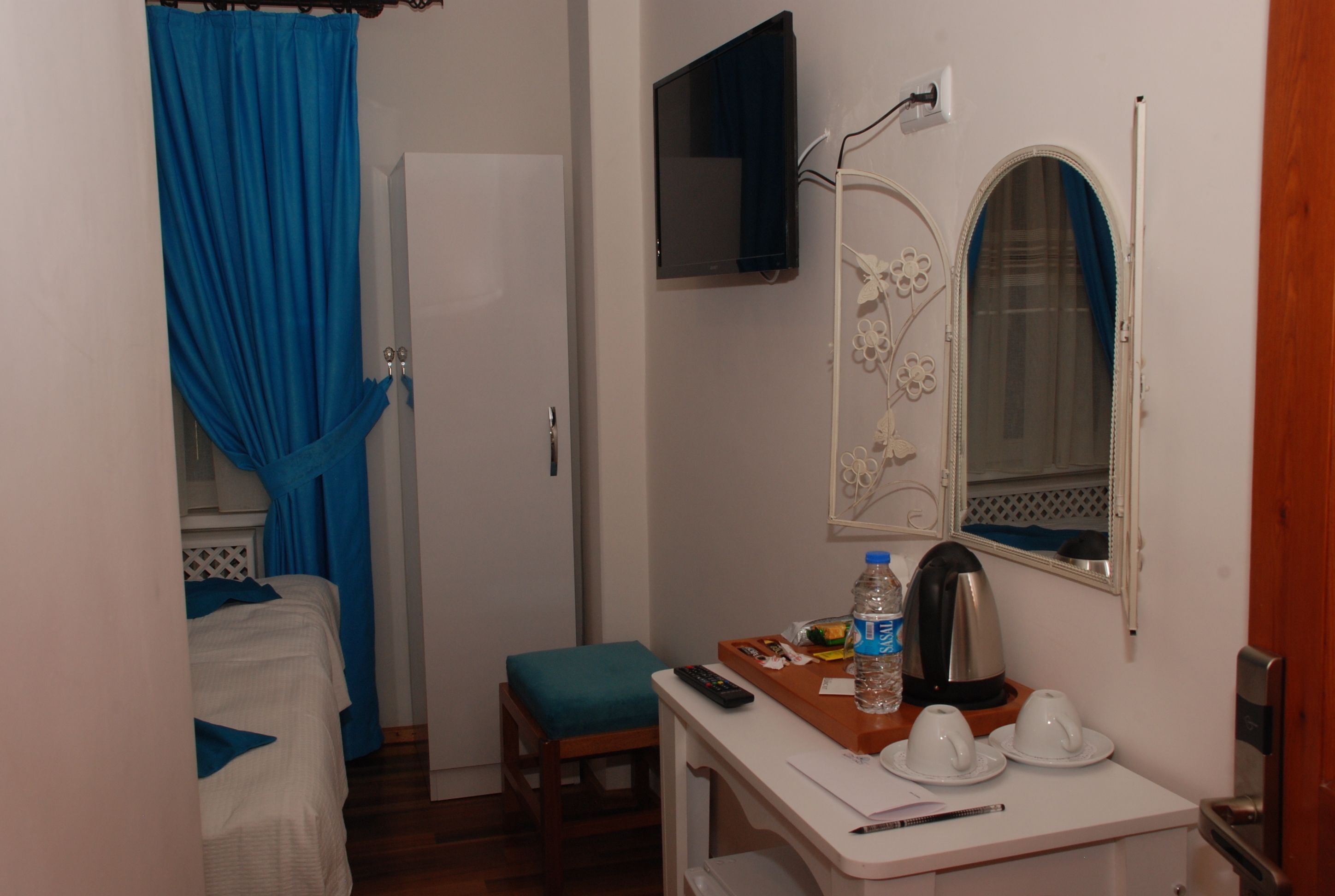 A hotel room with a bed, a small table with a remote control, a water bottle, and a mirror with a floral design.