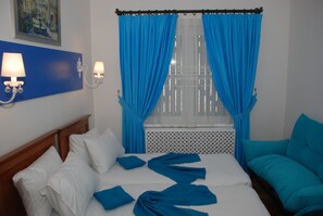 A hotel room with a bed, a window with blue curtains, a wall-mounted lamp, and a painting above the bed. - Mavi Inci Park Otel (Erdek)