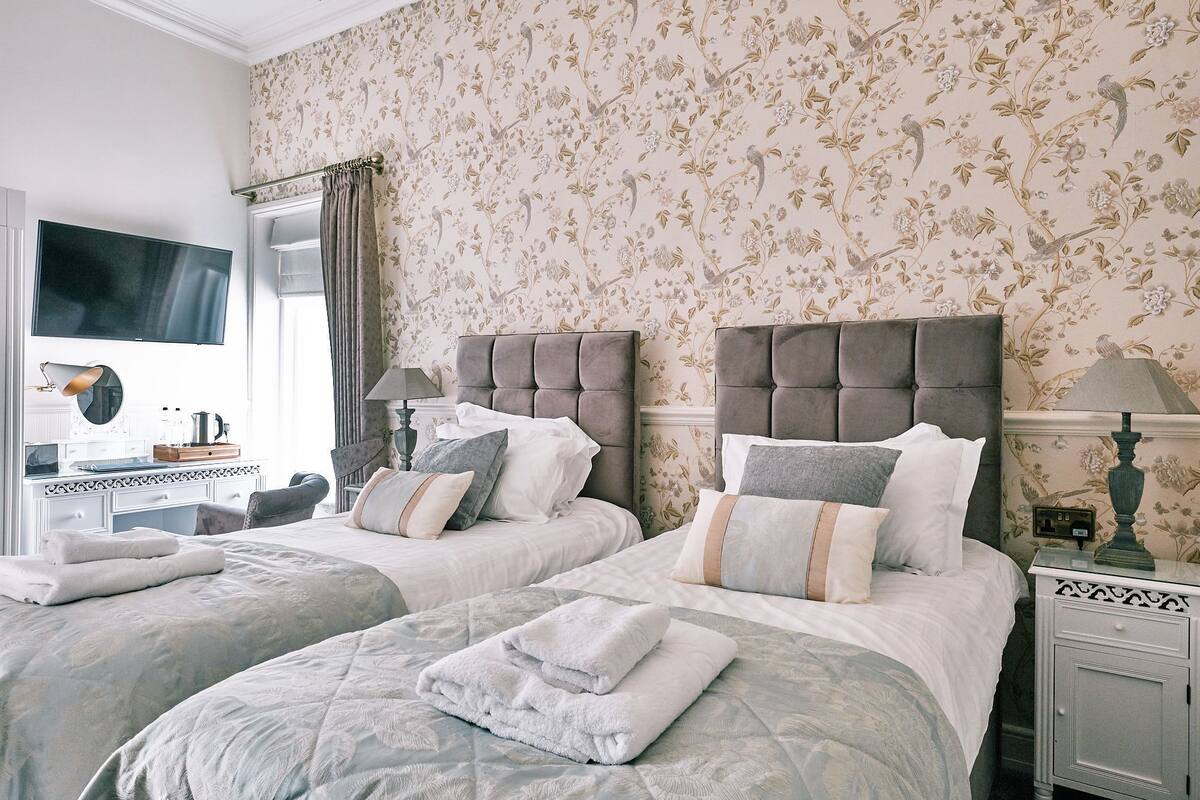 Habitación de hotel con dos camas, televisión, mesita de noche con lámpara y pared con estampado floral.