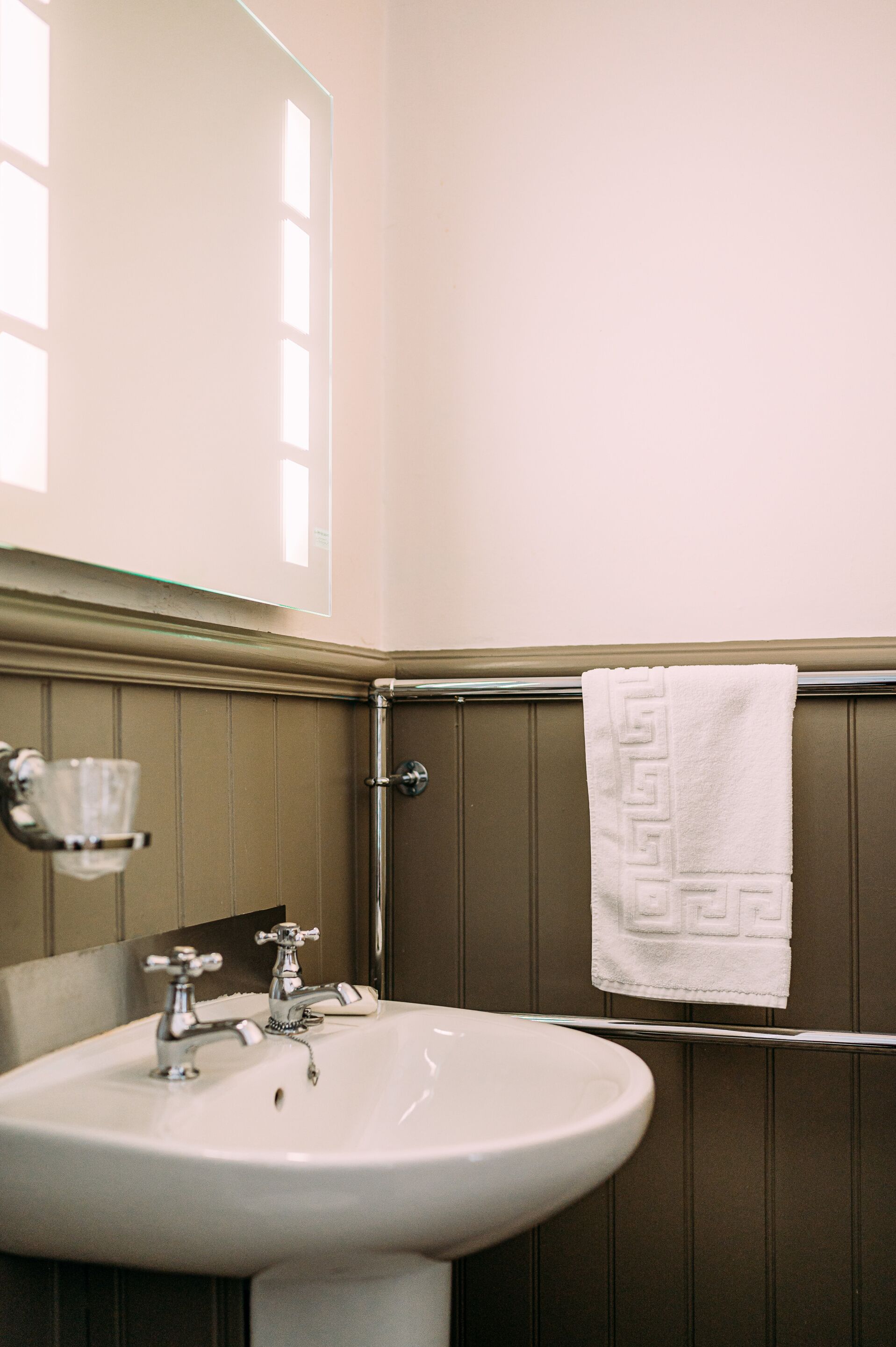 Un baño con un lavamanos blanco, dos grifos, un recipiente de vidrio a la izquierda y una toalla con estampado geométrico colgada en un toallero metálico.