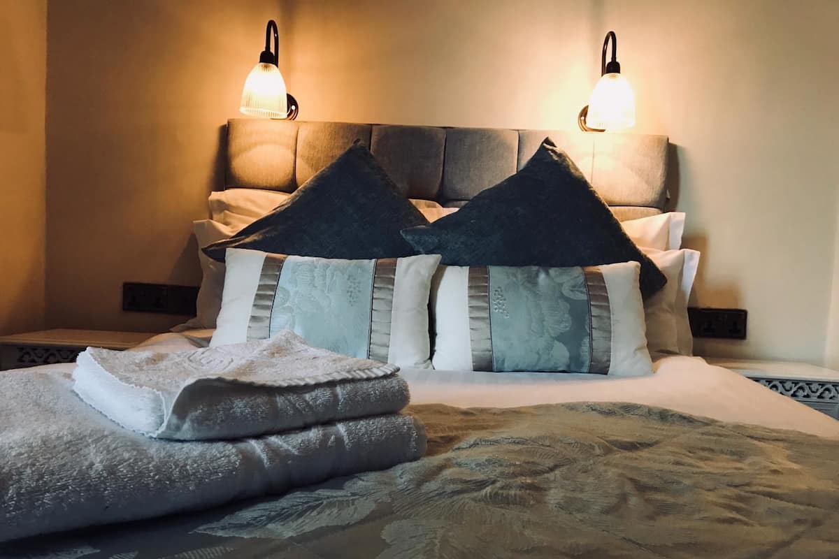 Una cama bien hecha con una colcha estampada, varios almohadones y dos lámparas fijadas a la pared.