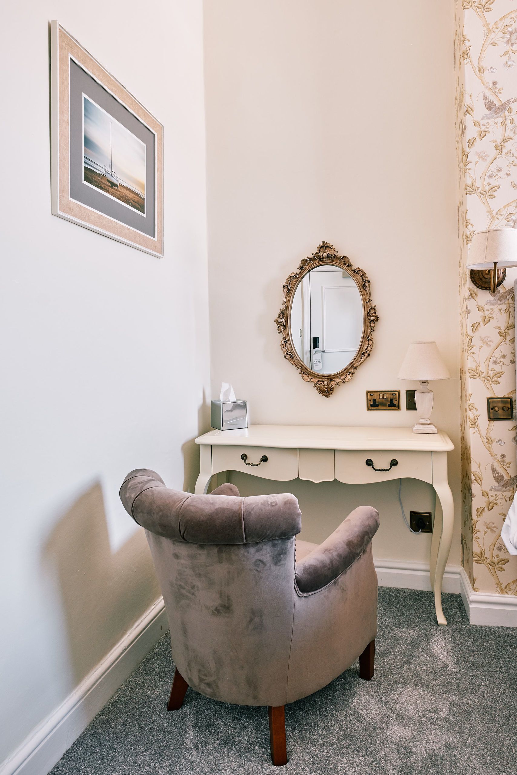 Une chambre d’angle avec une coiffeuse, un miroir, une chaise et un tableau encadré au mur.