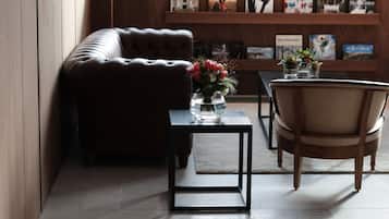 A modern living room with a leather sofa, a wooden armchair, a small black table with a vase of flowers, and a bookshelf with books and decorative items.