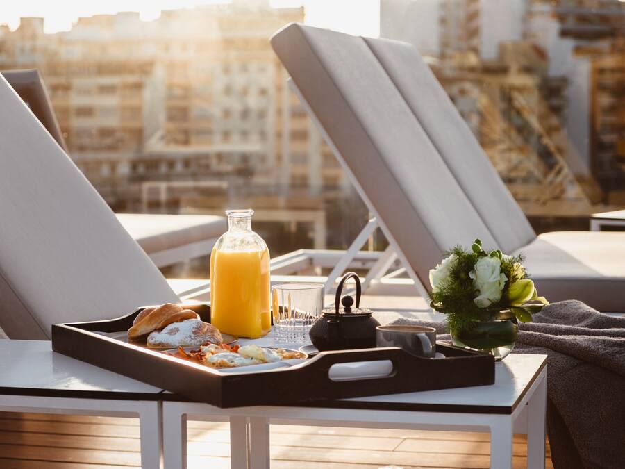 Una bandeja de desayuno con croissants, jugo de naranja y una tetera sobre una mesa con sillones y una vista a la ciudad al fondo.