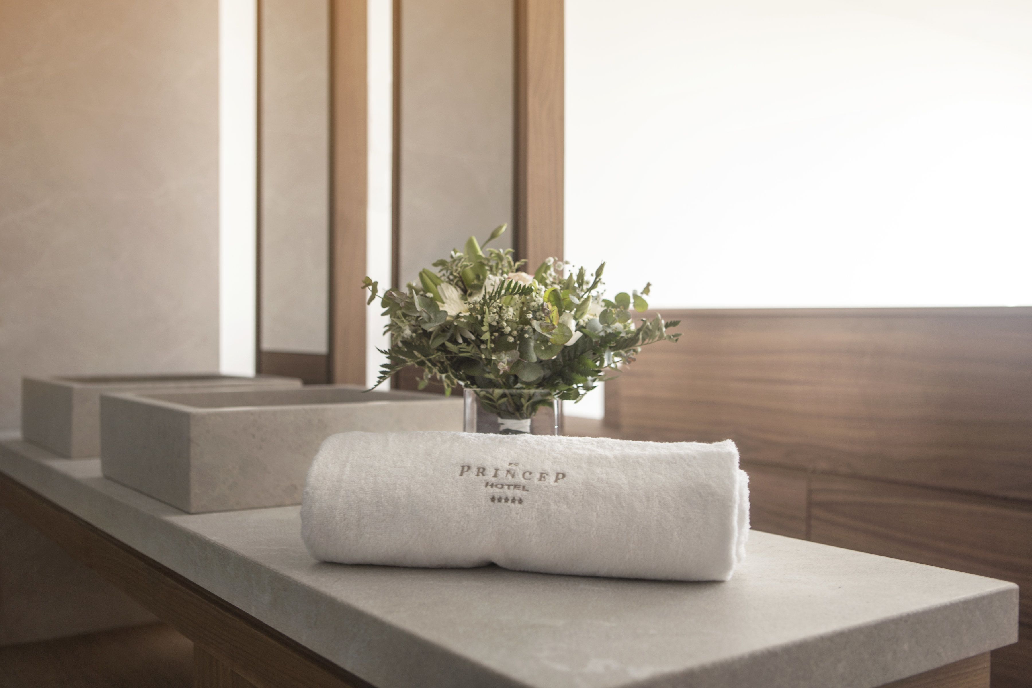 Een netjes opgevouwen witte handdoek met de tekst 'PRINCEP HOTEL' op een stenen rand, een kleine vaas met groene planten en een houten achtergrondpaneel.