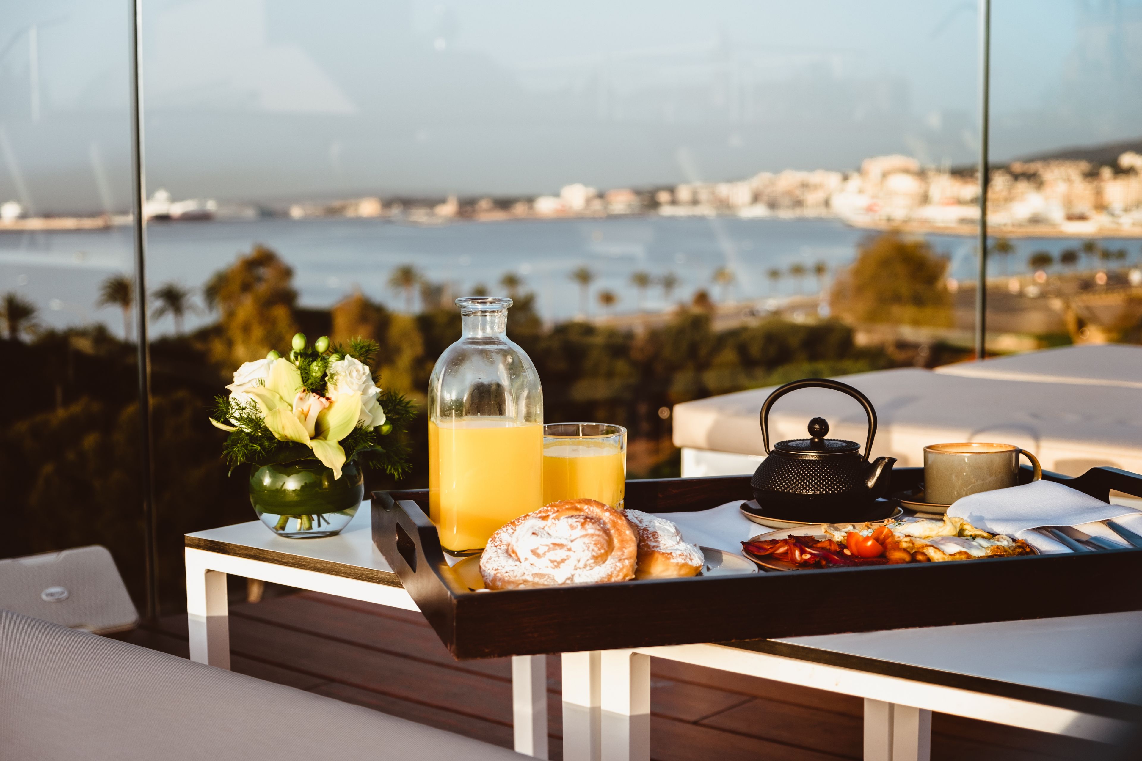 A breakfast tray with pastries, a teapot, a mug, and a glass of orange juice on a table with a view of the sea and cityscape.