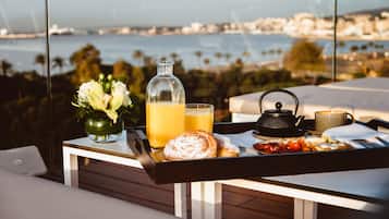 A breakfast tray with pastries, a teapot, a mug, and a glass of orange juice on a table with a view of the sea and cityscape.