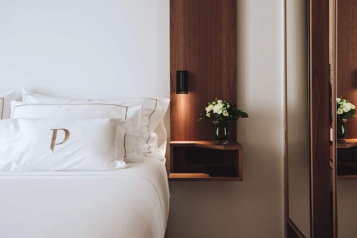 Una cama impecablemente arreglada con sábanas blancas y un cojín con la letra 'P' en dorado. Hay una mesita de noche de madera con un jarrón de flores blancas y una lámpara fijada a la pared.
