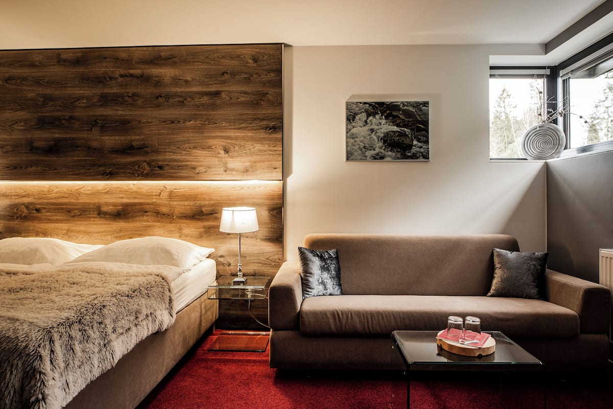 Ein modernes Hotelzimmer mit einem Holzkopfteil, einer Sitzecke, einem kleinen Tischchen mit Tablett und einem Fenster mit Blick auf Bäume.