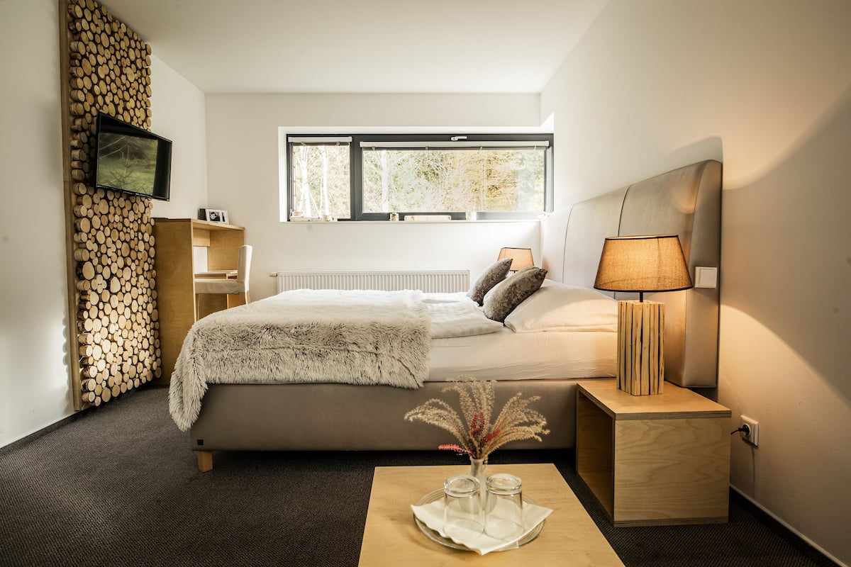 Ein modernes Schlafzimmer mit Bett, Nachttisch, Lampe und einem Fenster mit Blick auf Bäume.