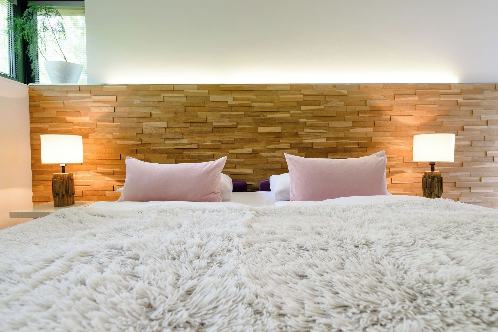A bedroom with a wooden headboard, two bedside lamps, a fluffy white bedspread, and a potted plant by the window.
