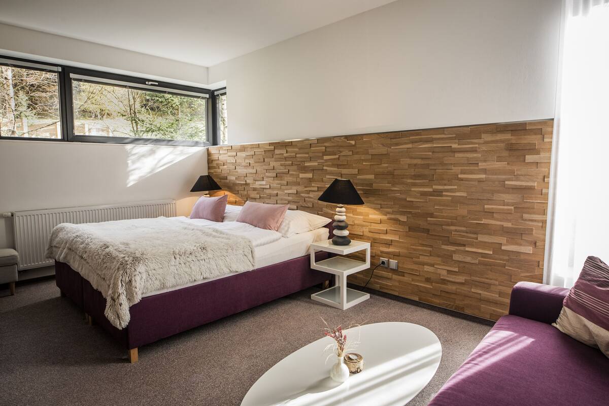 Ein modernes Schlafzimmer mit einem Bett, Nachttischen, einem kleinen weißen Tisch und Blick ins Freie durch ein Fenster.