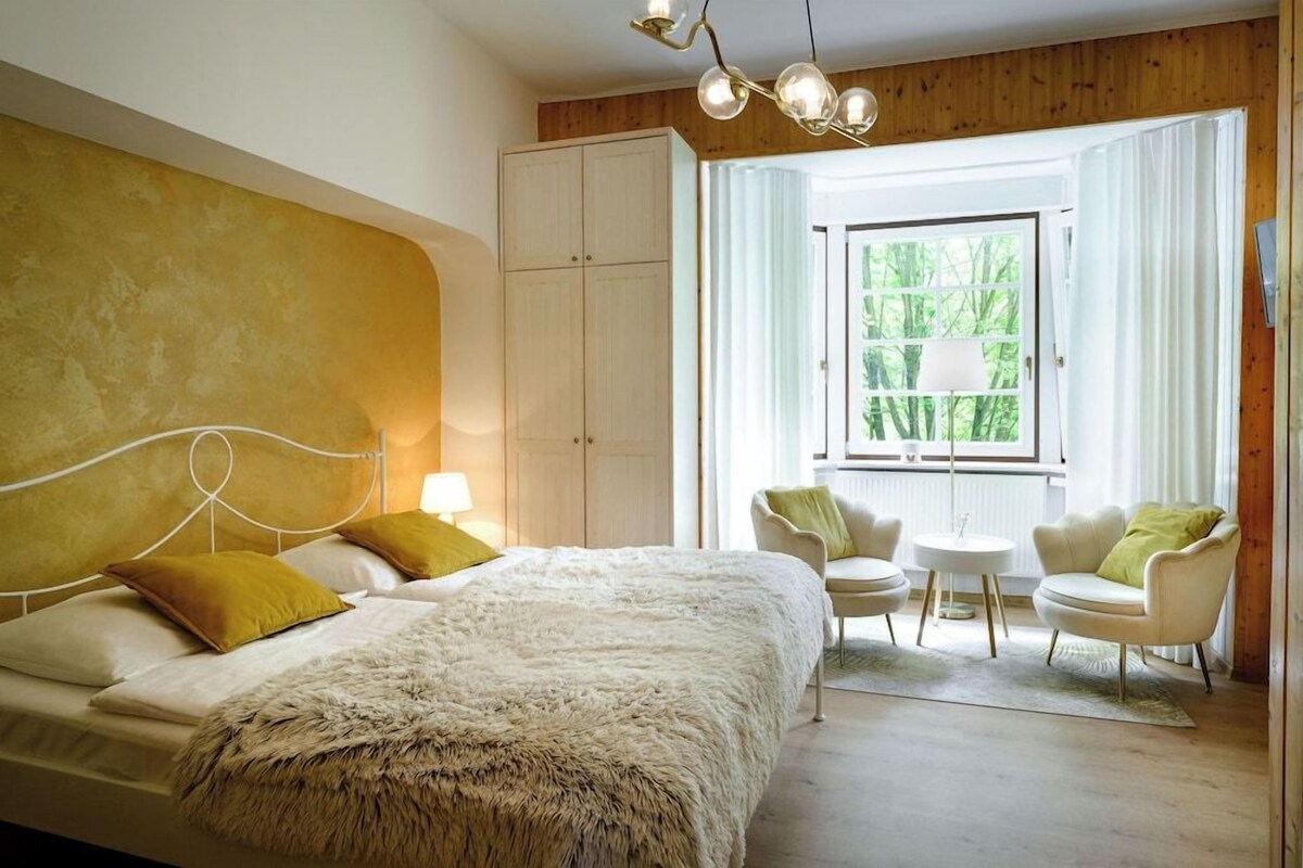 Ein Schlafzimmer mit einem großen Bett, einem Kleiderschrank, einem Fenster mit Vorhängen und einer Sitzecke mit zwei Stühlen und einem kleinen Tisch.
