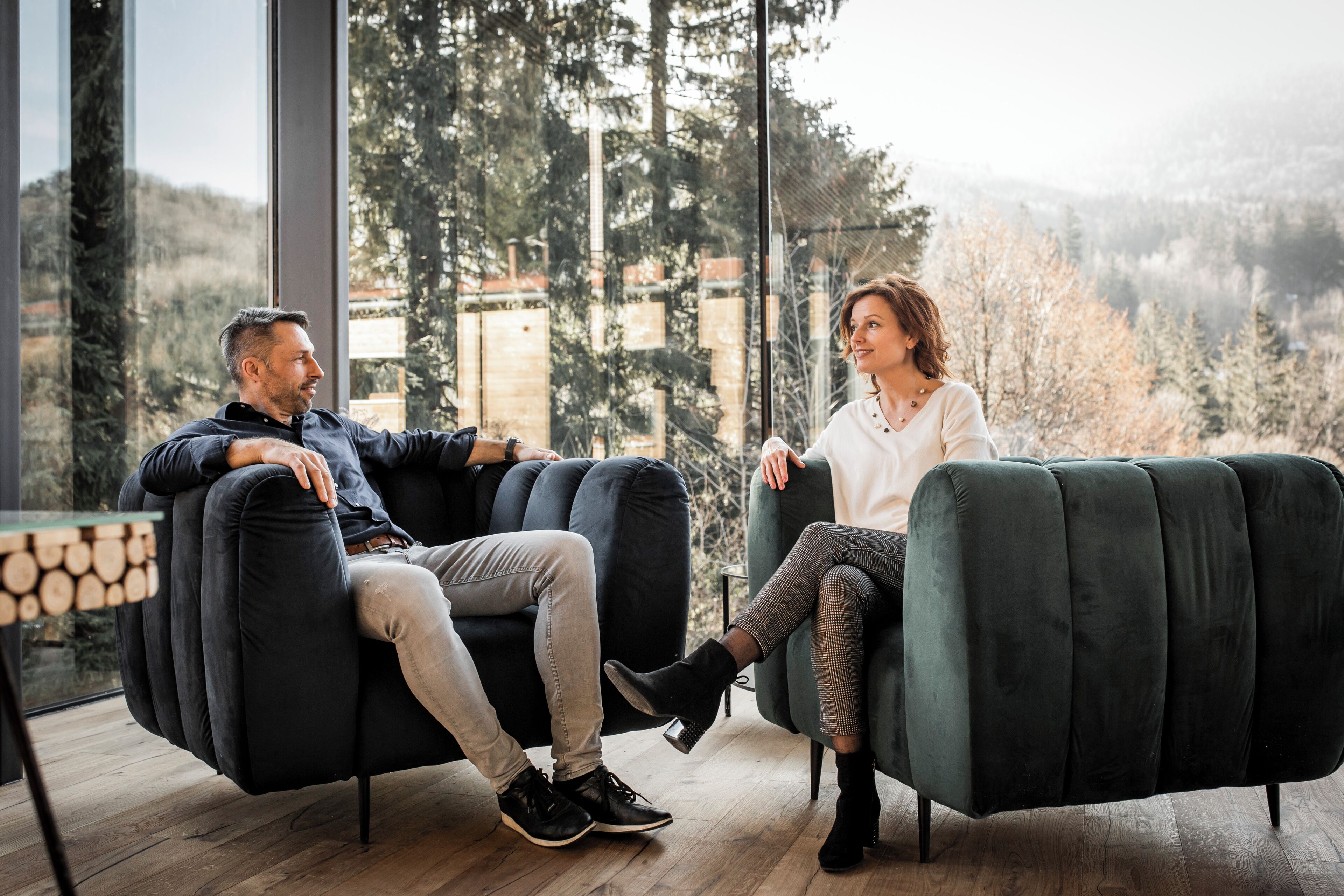Two people seated on dark sofas in a room with large windows overlooking a forested area.