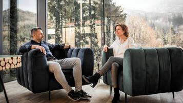 Two people seated on dark sofas in a room with large windows overlooking a forested area.