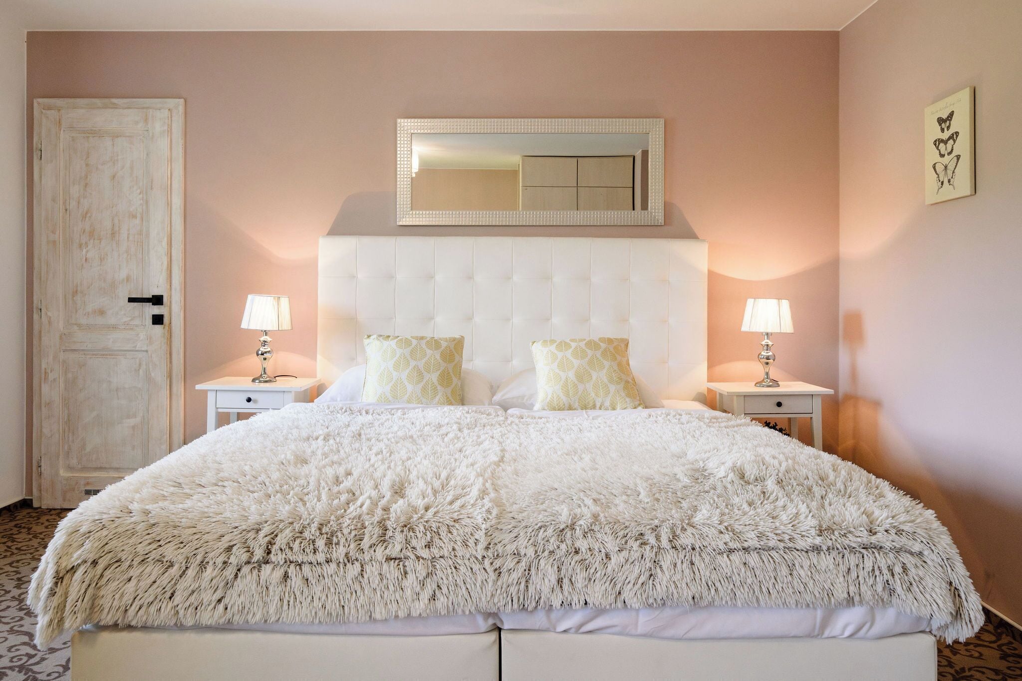 A bedroom with a tufted headboard, two bedside tables with lamps, a fluffy white bedspread, and a large mirror above the headboard.