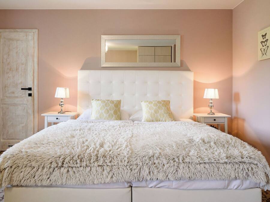 A bedroom with a tufted headboard, two bedside tables with lamps, a fluffy white bedspread, and a large mirror above the headboard.
