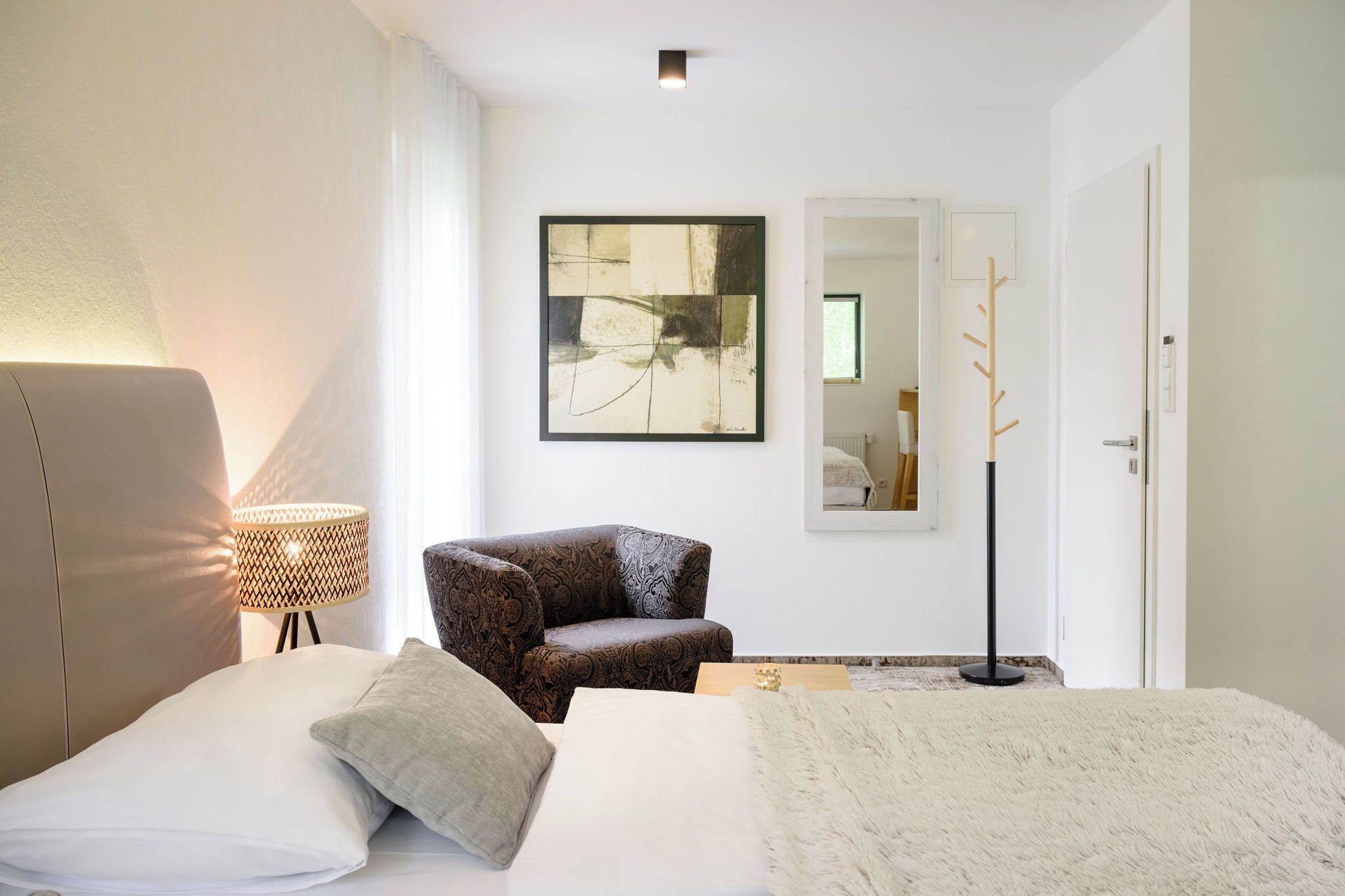 A modern hotel room with a bed, a patterned armchair, a floor lamp, and a large abstract painting on the wall.