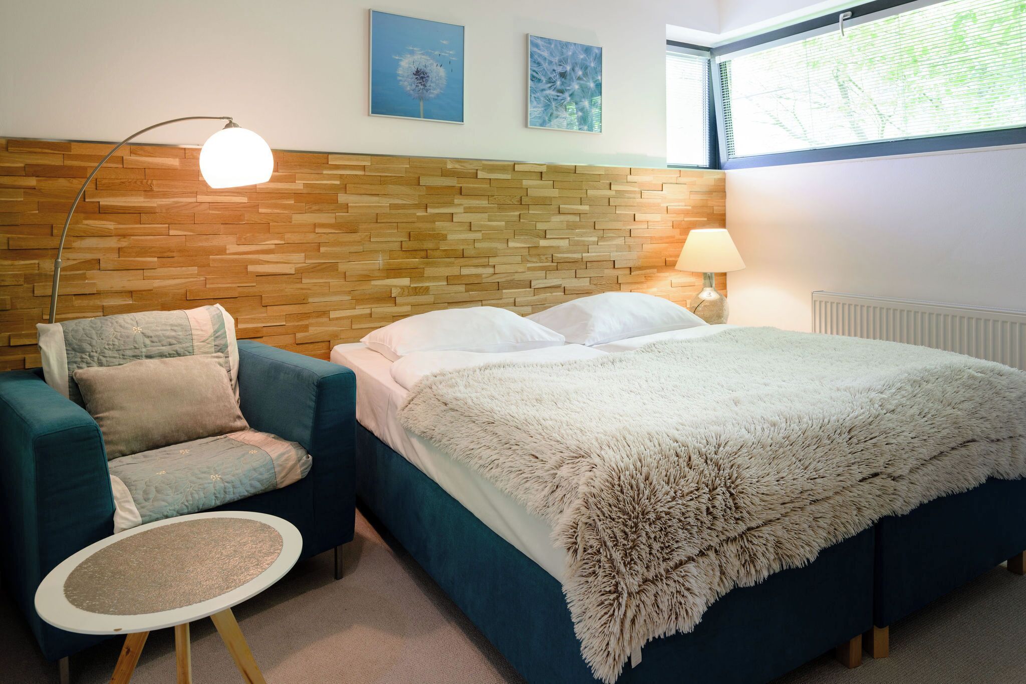 A bedroom with a wooden headboard, a bed with a fluffy white blanket, a blue armchair, a small round table, and two wall-mounted lamps.