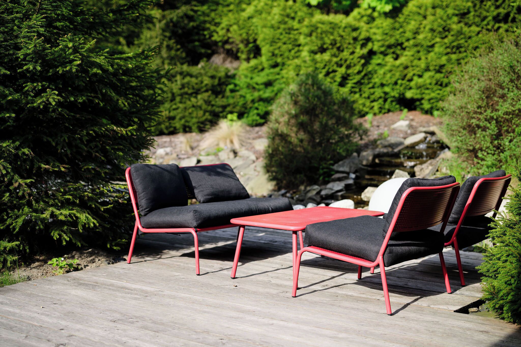 Eine Terrasse mit Gartenmöbeln, darunter eine Sitzecke und Stühle, umgeben von viel Grün und einem kleinen Wasserfall.